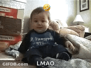 A Baby Wearing A Varsity Byg Shirt Is Sitting On A Bed GIF