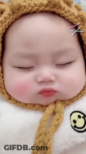 A Baby Wearing A Yellow Knitted Hat And A White Bib With A Smiley Face On It . GIF