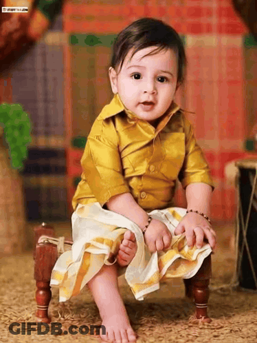 A Baby Wearing A Yellow Shirt And White Skirt Is Sitting On A Small Wooden Chair GIF