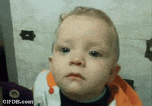 A Baby With A Sad Look On His Face Is Wearing An Orange Sweater GIF