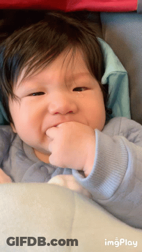 A Baby With A Tear Coming Out Of His Eye Is Sitting In A Stroller With A Blue Sweater On GIF