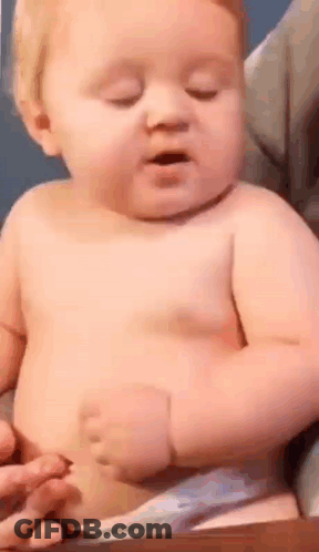 A Baby Without A Shirt Is Sitting On A Table With His Eyes Closed And His Mouth Open . GIF