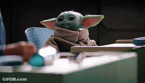 A Baby Yoda Is Sitting At A Table With A Scarf Around His Neck . GIF