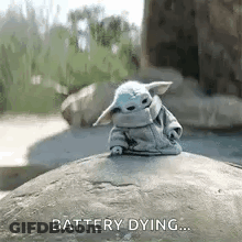 A Baby Yoda Is Sitting On Top Of A Rock With The Words `` Battery Dying '' Written On It . GIF