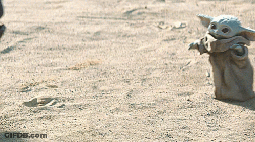 A Baby Yoda Toy Is Standing In The Sand Looking At Something GIF