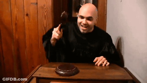 A Bald Man In A Black Robe Is Sitting At A Wooden Table With A Hammer In His Hand GIF