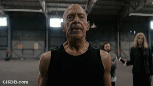 A Bald Man In A Black Tank Top Is Standing In A Dark Room GIF