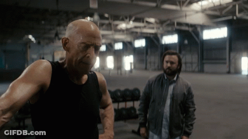 A Bald Man In A Black Tank Top Stands Next To Another Man In A Grey Jacket GIF