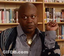 A Bald Man In A Blue Shirt And Striped Sweater Is Standing In Front Of A Bookshelf . GIF