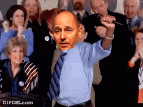 A Bald Man In A Blue Shirt And Tie Holds A Microphone In Front Of A Crowd GIF