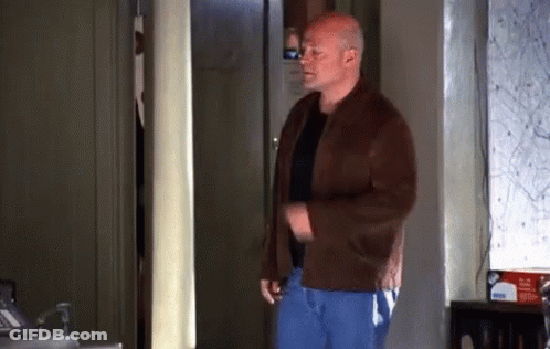 A Bald Man In A Brown Jacket And Blue Pants Is Standing In A Room . GIF