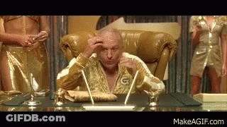 A Bald Man In A Gold Robe Is Sitting At A Desk With A Pen In His Hand . GIF