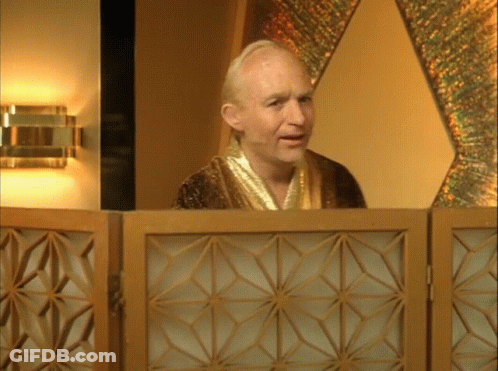 A Bald Man In A Gold Robe Stands Behind A Wooden Screen GIF