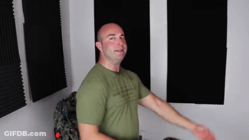 A Bald Man In A Green T-Shirt Is Standing In Front Of A Wall With Foam Panels . GIF