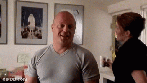 A Bald Man In A Grey Shirt Is Smiling While A Woman Stands Behind Him GIF