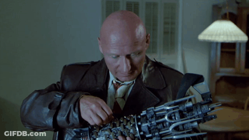 A Bald Man In A Leather Jacket And Tie Is Holding A Mechanical Arm GIF