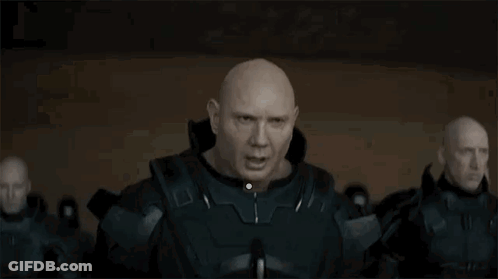 A Bald Man In A Military Uniform Is Standing In Front Of A Group Of Bald Men In A Dark Room . GIF