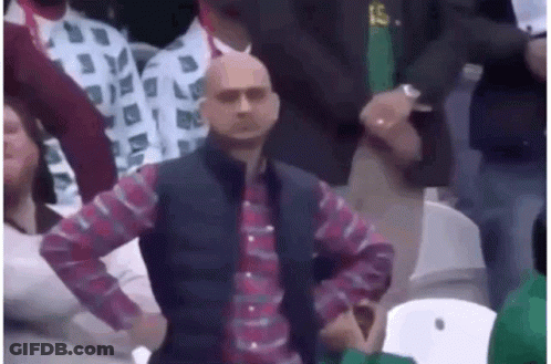 A Bald Man In A Plaid Shirt And Vest Is Sitting In A Crowd . GIF