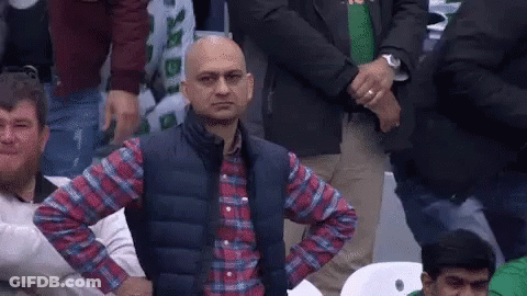 A Bald Man In A Plaid Shirt And Vest Is Sitting In A Crowd With His Hands On His Hips . GIF