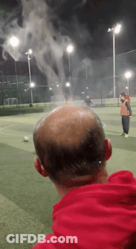 A Bald Man In A Red Hoodie Is Watching A Soccer Game GIF