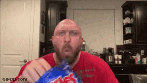 A Bald Man In A Red Shirt Is Eating A Blue Bag Of Chips . GIF