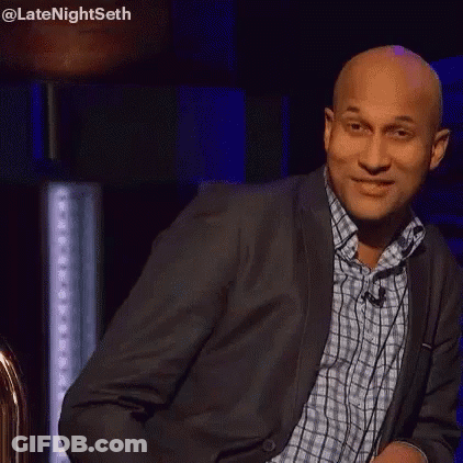 A Bald Man In A Suit And Plaid Shirt Is Standing In Front Of A Microphone And Making A Funny Face . GIF