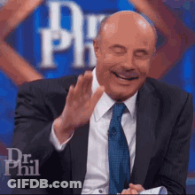 A Bald Man In A Suit And Tie Is Laughing And Waving His Hand . GIF