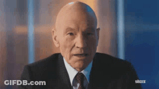 A Bald Man In A Suit And Tie Is Sitting In Front Of A Blue Wall . GIF