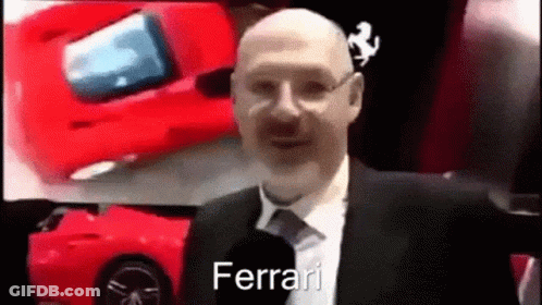 A Bald Man In A Suit And Tie Is Talking Into A Microphone While Standing In Front Of A Red Ferrari . GIF
