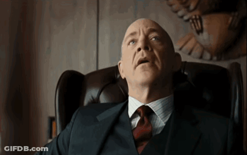A Bald Man In A Suit And Tie Sits In A Chair GIF