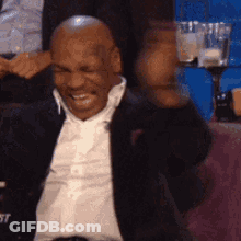 A Bald Man In A Suit And White Shirt Is Sitting In A Chair With His Fist In The Air . GIF