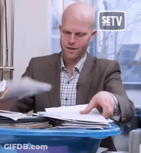 A Bald Man In A Suit Is Sitting At A Desk With Papers In A Blue Bin . GIF