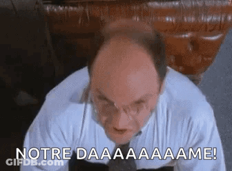 A Bald Man In A White Shirt And Tie Is Sitting In Front Of A Table And Making A Funny Face . GIF