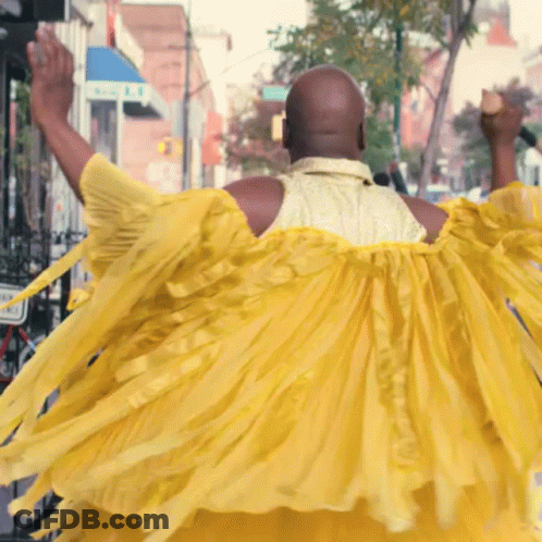 A Bald Man In A Yellow Dress Is Walking Down A Street GIF