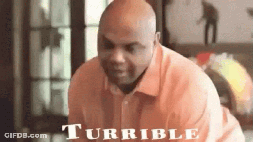 A Bald Man In An Orange Shirt Is Sitting On A Couch And Looking At The Camera . GIF