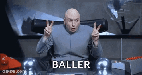A Bald Man Is Giving A Peace Sign While Sitting At A Desk With The Word Baller Written On It . GIF