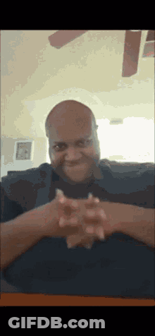 A Bald Man Is Sitting At A Table With His Hands Folded GIF