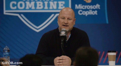 A Bald Man Is Speaking Into A Microphone In Front Of A Sign That Says Combine GIF