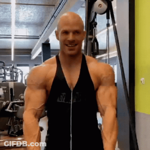 A Bald Man Is Standing In A Gym Wearing Headphones And A Black Tank Top . GIF