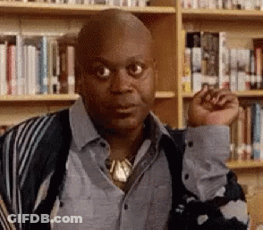 A Bald Man Is Standing In Front Of A Bookshelf In A Library With His Fist In The Air . GIF