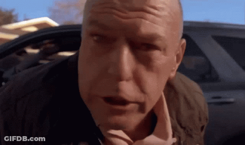 A Bald Man Is Standing In Front Of A Car And Looking At The Camera . GIF