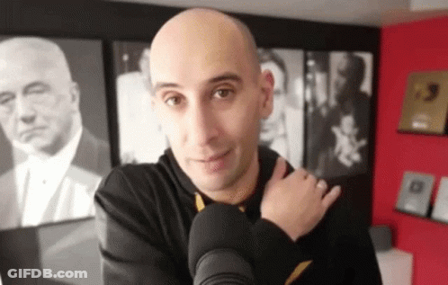 A Bald Man Is Standing In Front Of A Microphone In A Room With Pictures On The Wall Behind Him . GIF