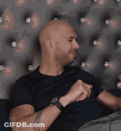 A Bald Man Wearing A Black Shirt And A Watch Is Sitting On A Couch GIF