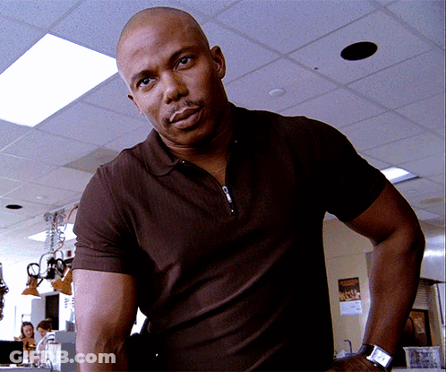 A Bald Man Wearing A Brown Polo Shirt And A Watch Stands In A Room GIF