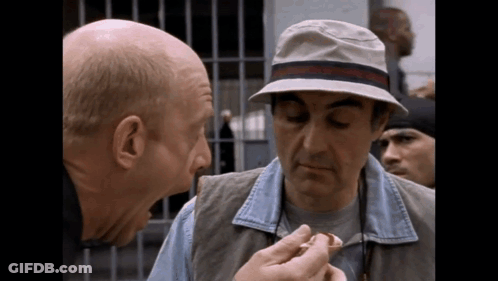 A Bald Man Wearing A Hat Is Talking To Another Man GIF