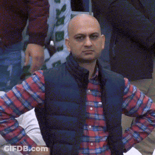 A Bald Man Wearing A Plaid Shirt And A Blue Vest Is Looking At The Camera With His Hands On His Hips GIF