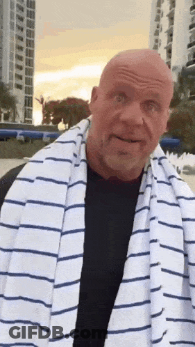 A Bald Man Wearing A Striped Towel Around His Neck Is Standing In Front Of A Building . GIF