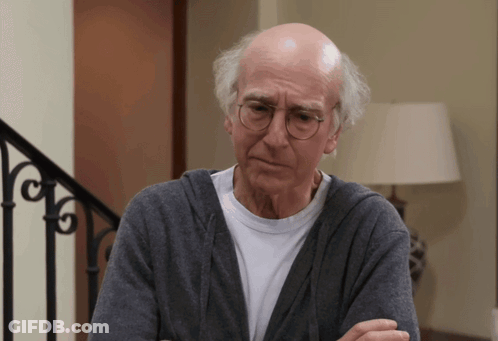 A Bald Man Wearing Glasses And A Gray Sweater Looks At The Camera GIF