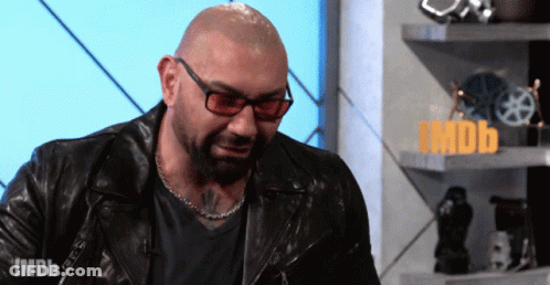 A Bald Man Wearing Glasses And A Leather Jacket Stands In Front Of A Sign That Says Imdb On It GIF