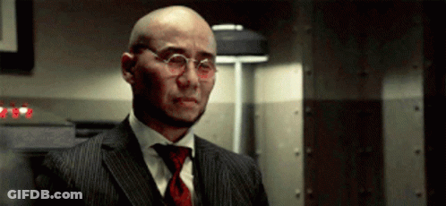A Bald Man Wearing Glasses And A Suit Stands In A Dark Room GIF